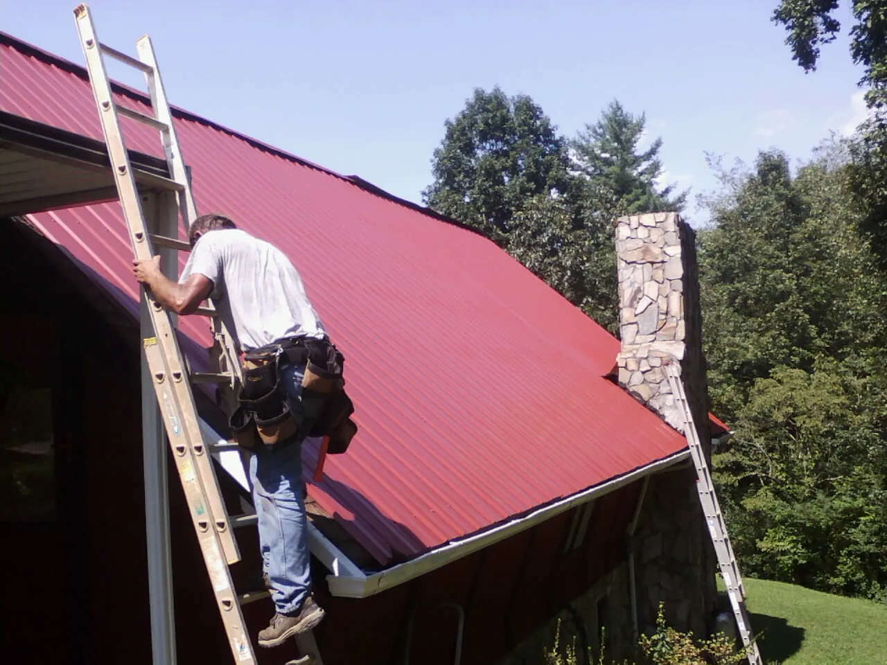 Roofer on a ladder beside a red metal roof and stone chimney — Soffit & Fascia Repair by Roofing Brothers in Newtown Square